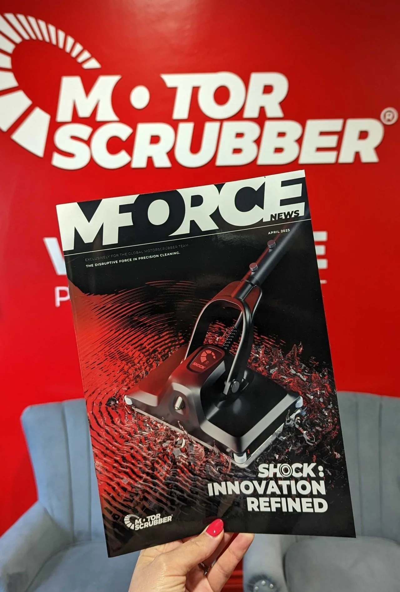 A hand holding a copy of MForce News with a scrubber render on the cover, in front of a MotorScrubber banner